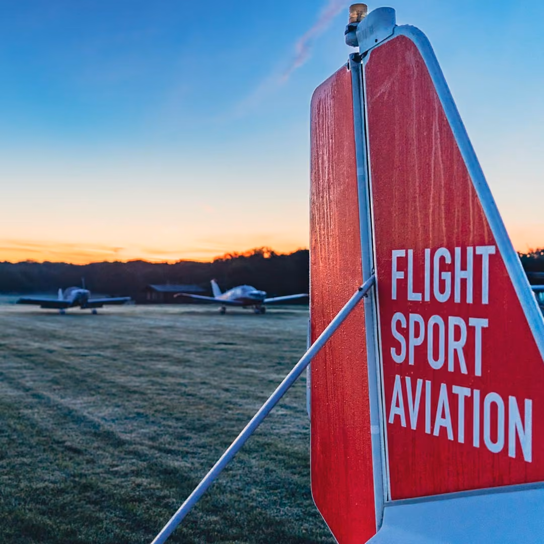 A red airplane tail with "Flight Sport Aviation" stands near two small planes at sunrise.