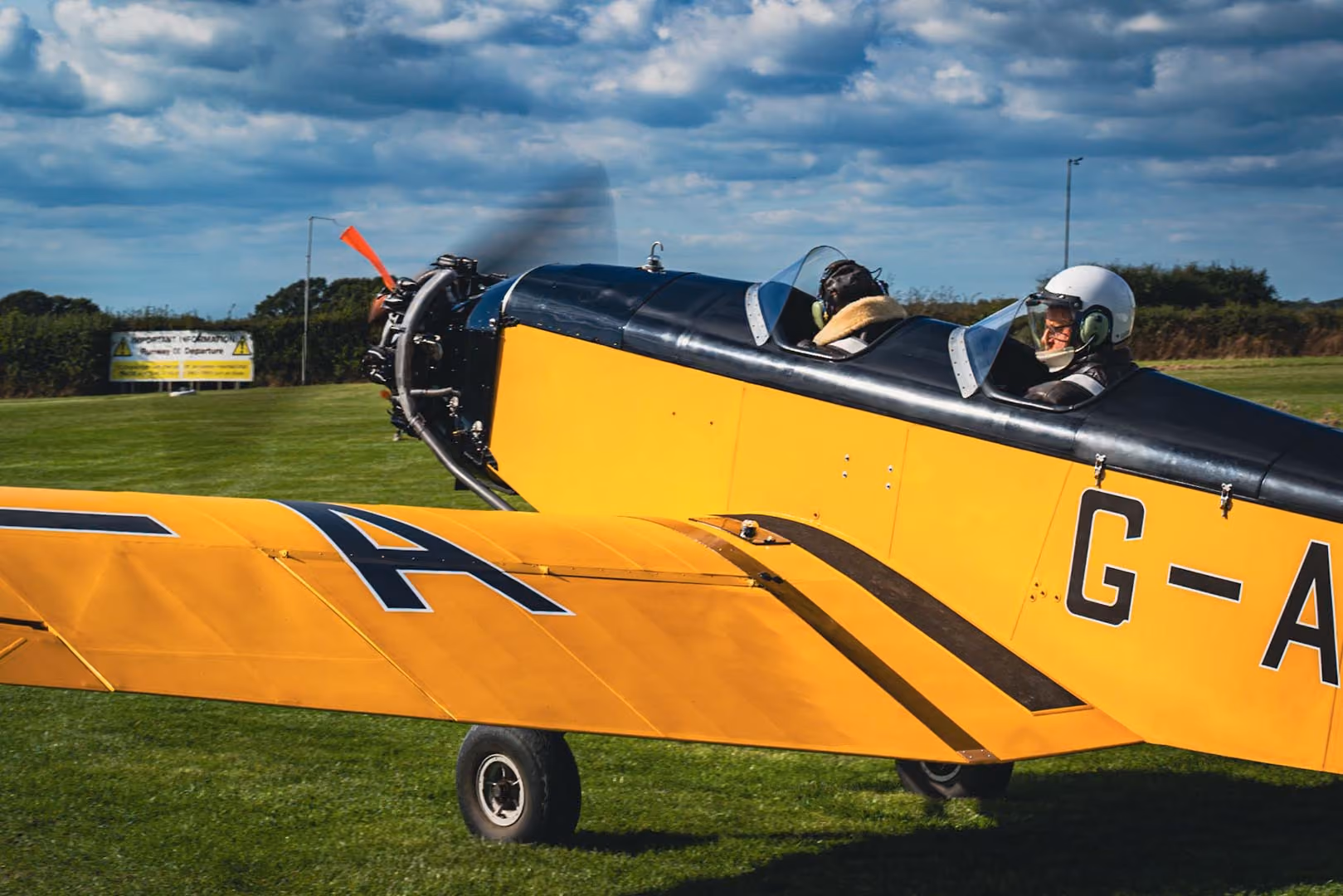 A yellow vintage airplane with two people inside is preparing for takeoff on a grassy field.