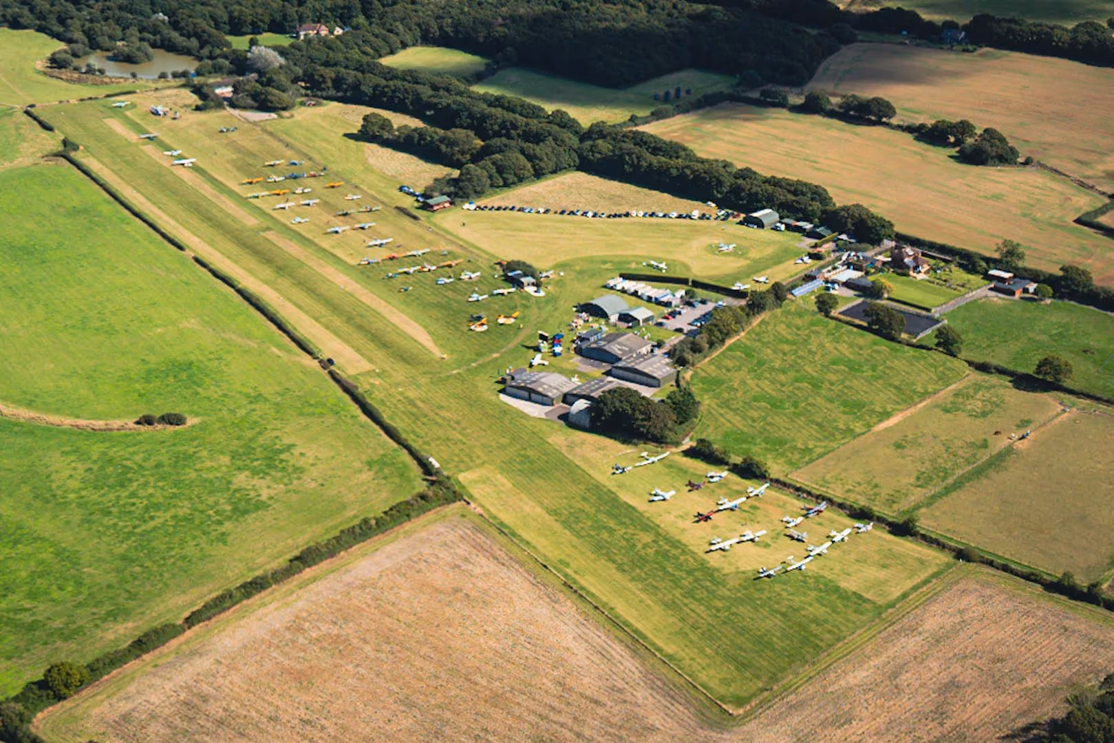 Aerial view of an airfield with numerous small airplanes parked on grassy runways and surrounding fields.