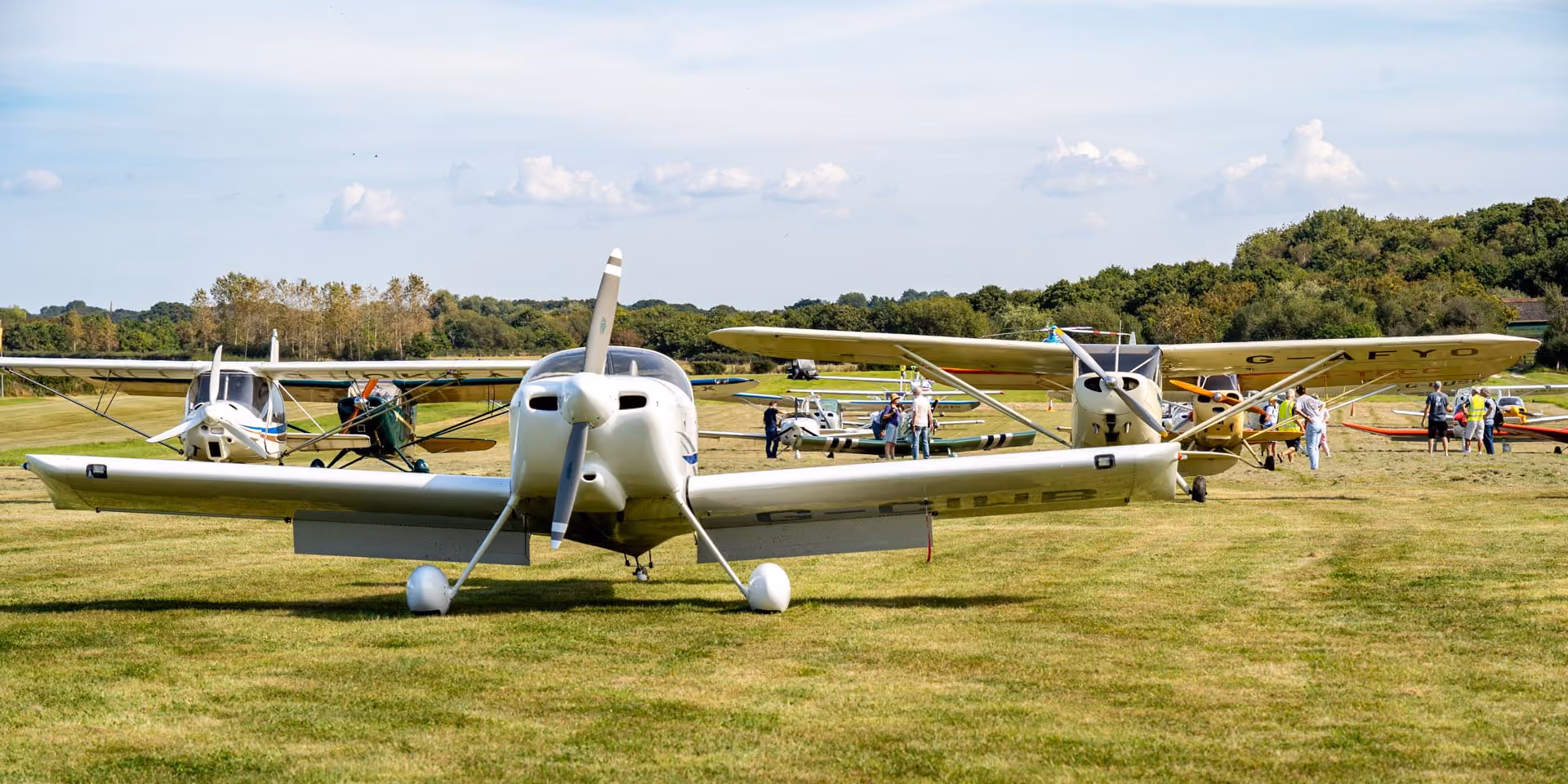 Several small airplanes parked on a grassy field with people walking around them.