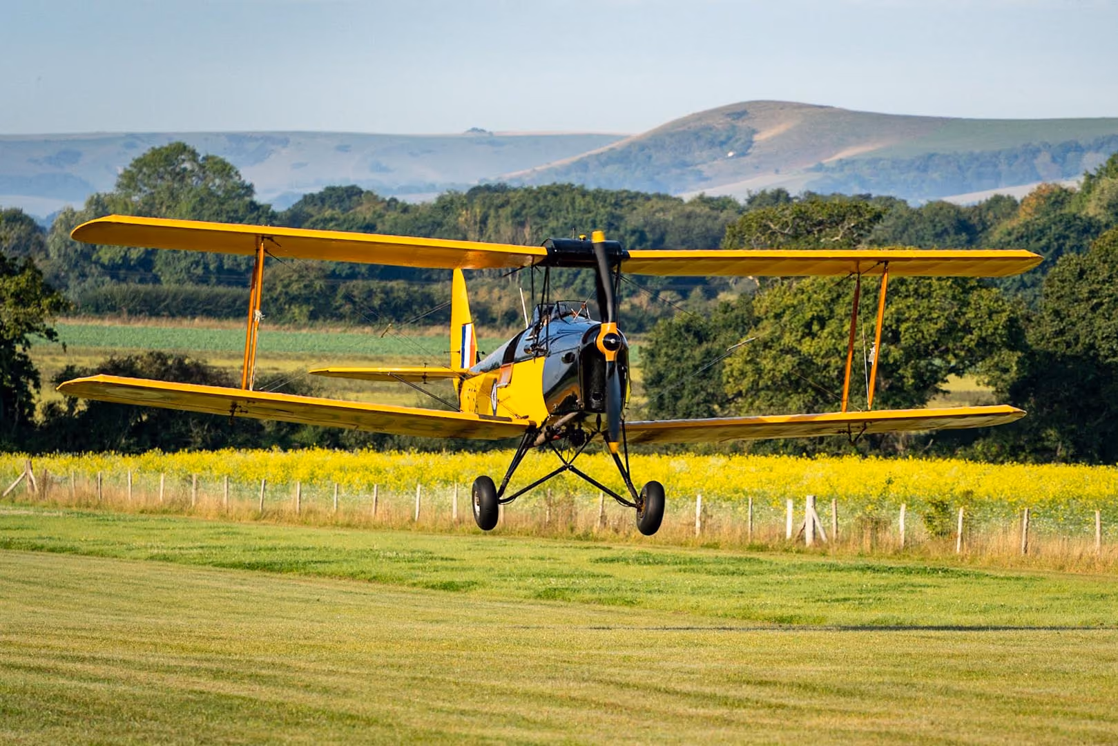 A yellow biplane is taking off from a grassy field surrounded by trees and hills.