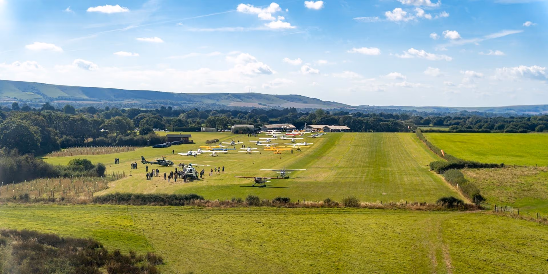 A small airfield with several parked light aircraft, surrounded by grassy fields and distant hills.