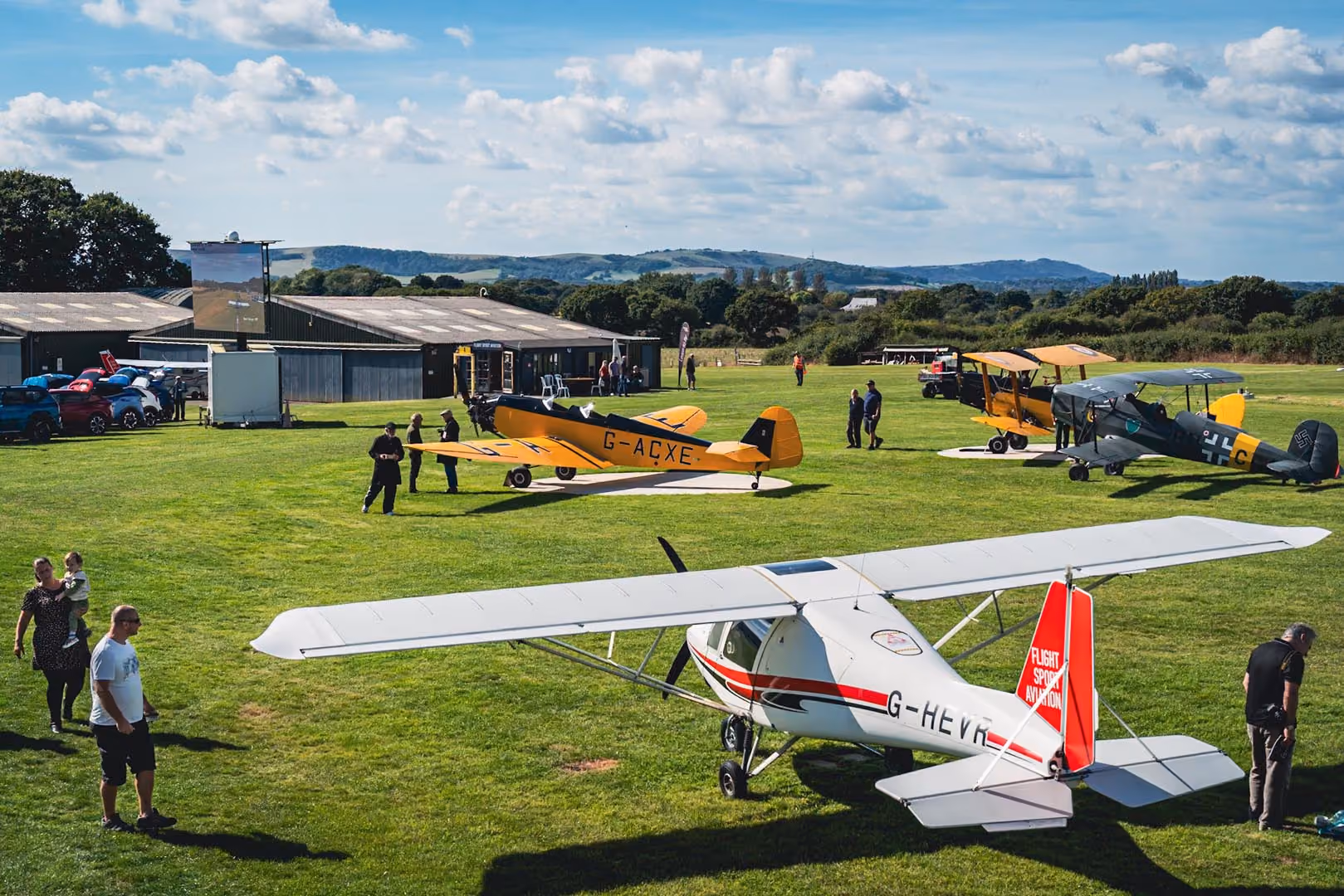 People stand near several vintage airplanes parked on a grassy field under a clear sky.