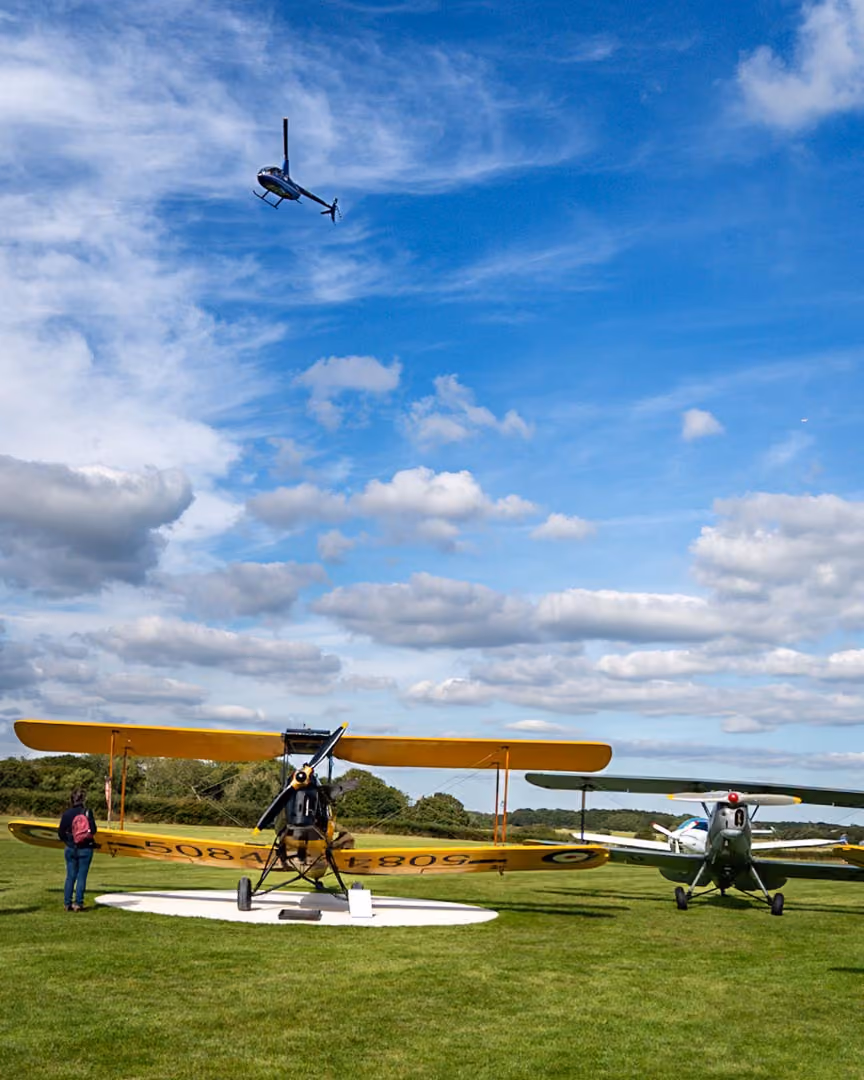 A person stands next to a yellow biplane on grassy field as a helicopter flies above.