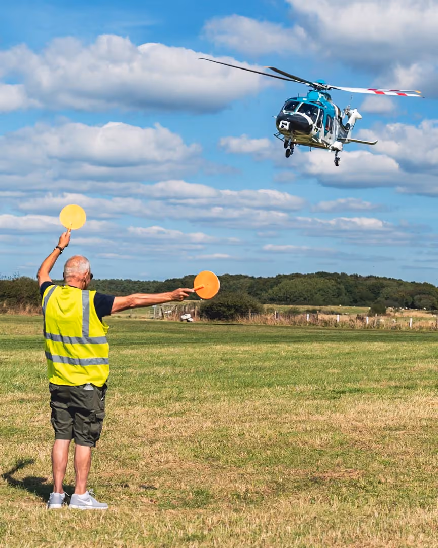 Man in safety vest directs helicopter landing using yellow paddles in open field.