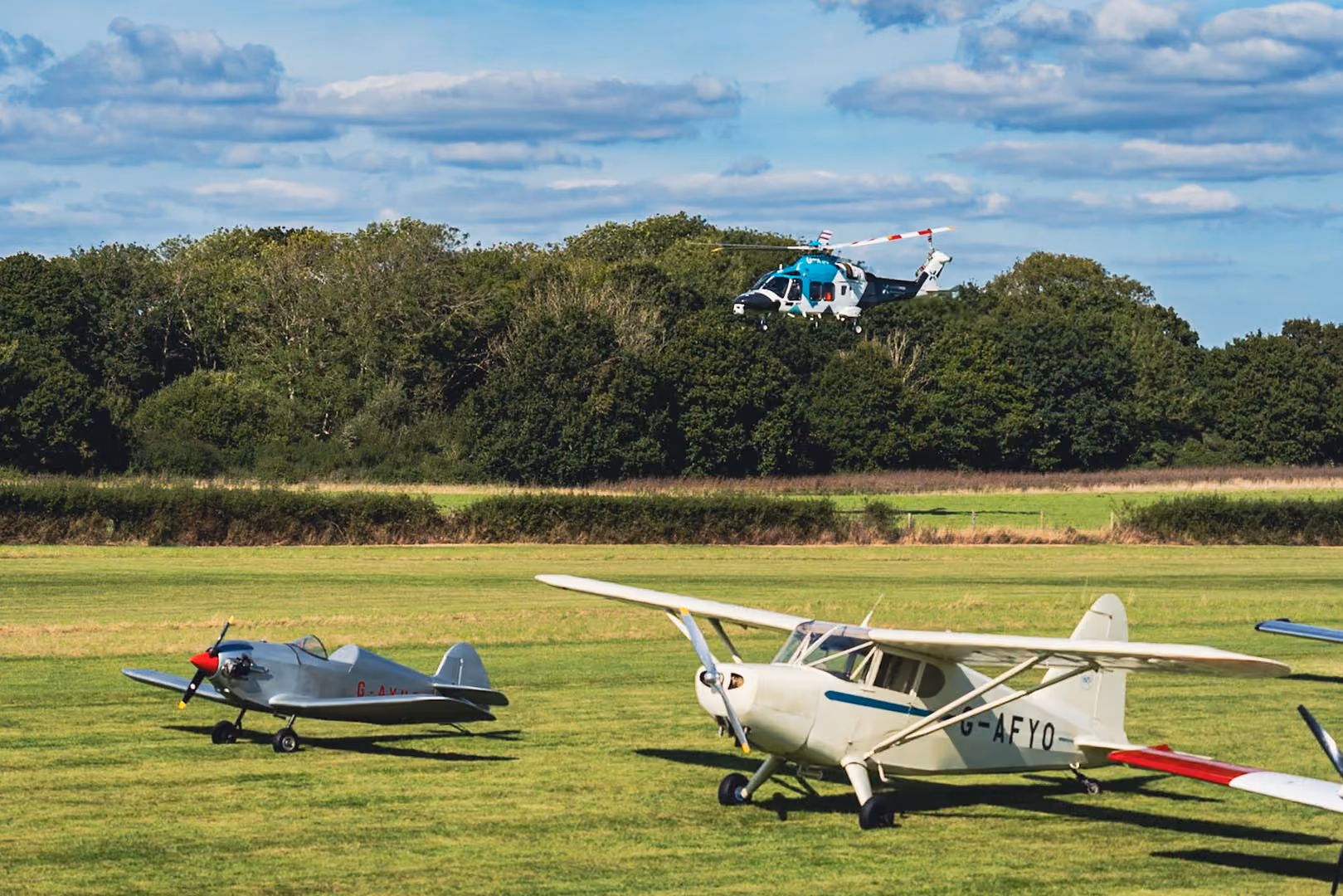 A helicopter flies over two small airplanes parked on a grassy field.