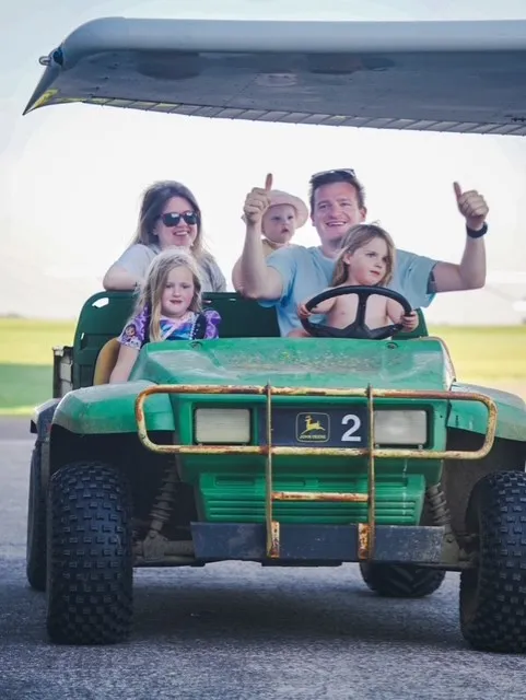A family of five rides in a green utility vehicle, with the driver giving thumbs up.