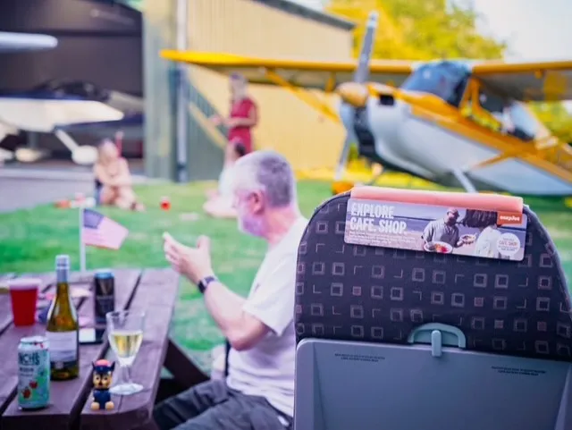 A person sits at an outdoor picnic table near small airplanes, with drinks on the table.