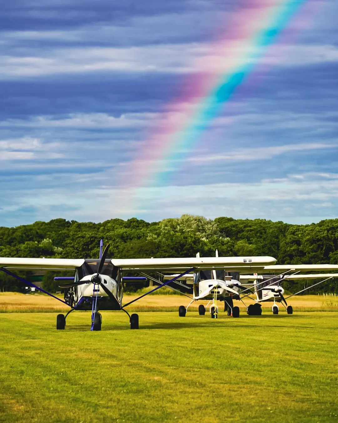 Three small airplanes parked on a grassy field with a rainbow in the sky above.