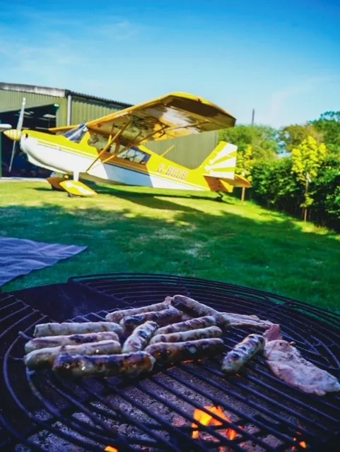 Sausages grilling on a barbecue with a small airplane parked nearby on the grass.