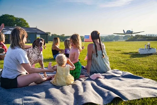 Several people and a dog sit on a blanket as a small plane flies by.