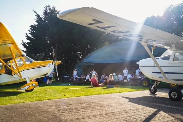 Two small airplanes are parked on grassy field, with people gathered under a canopy nearby.