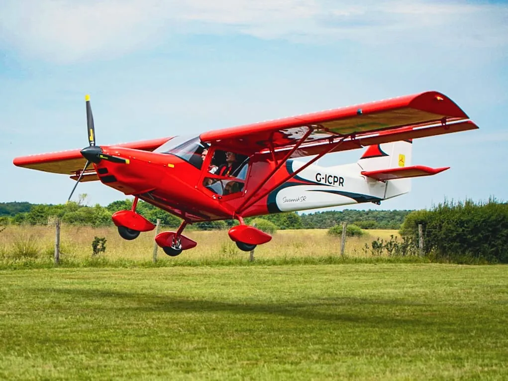 A red airplane is taking off from a grassy field under a clear sky.