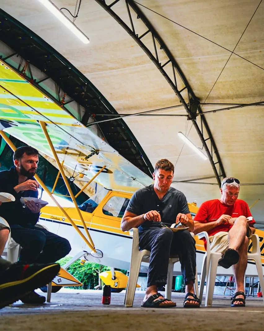Three men sit in chairs eating, with a yellow airplane parked nearby inside a hangar.