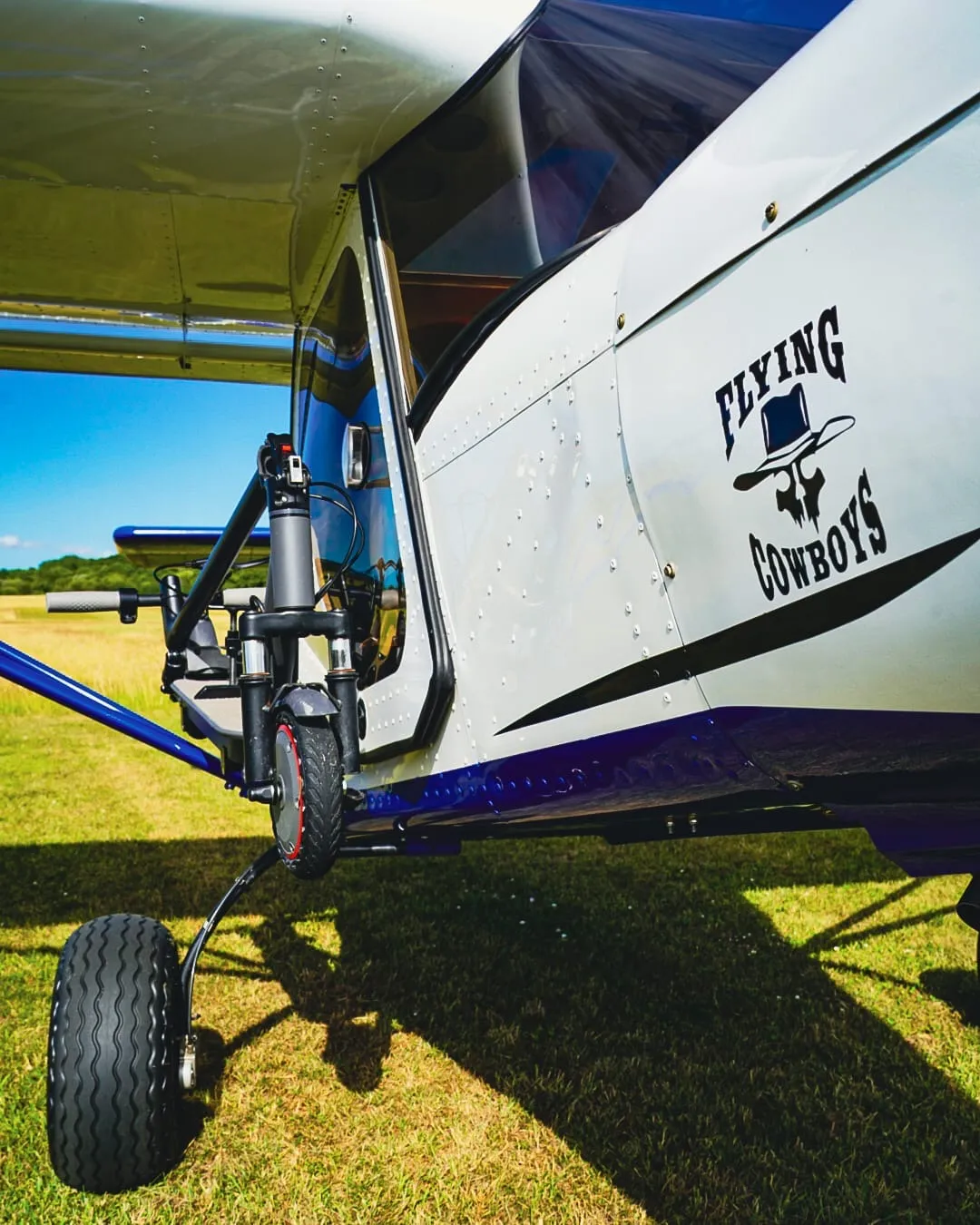 Small airplane with "Flying Cowboys" logo on side, parked on grassy field, wheel visible.