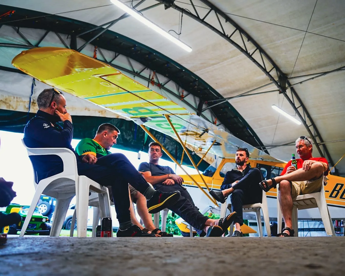 Five people sitting in a circle under an aircraft hangar, engaging in a discussion.