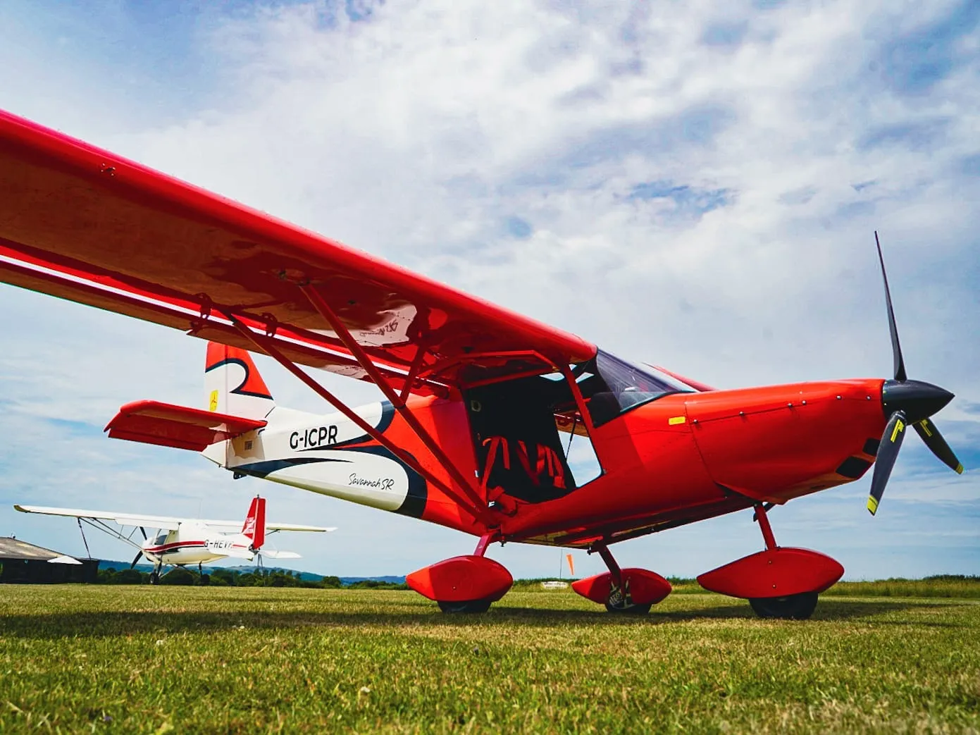 A bright red small airplane is parked on grassy ground under a cloudy sky.