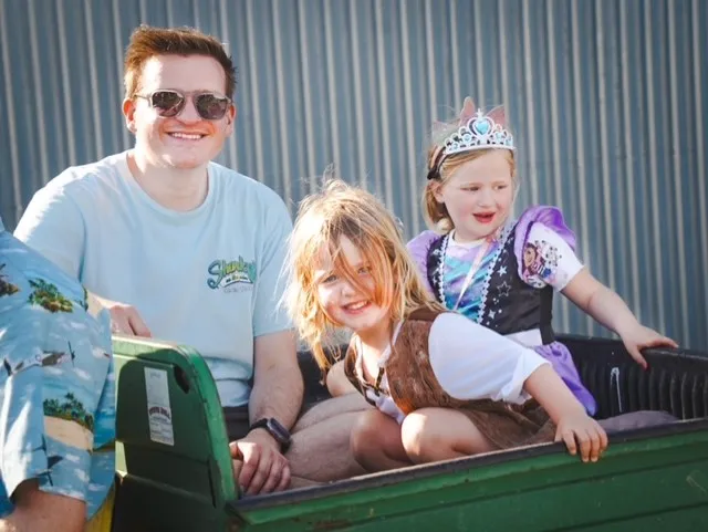 Man smiles while sitting with two children in a cart. One child wears a princess costume.