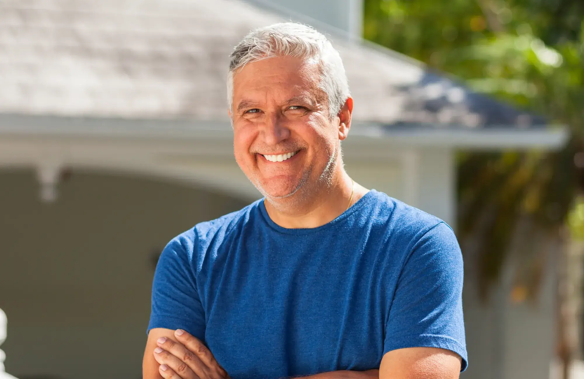 A smiling man with his arms crossed in front of a house.