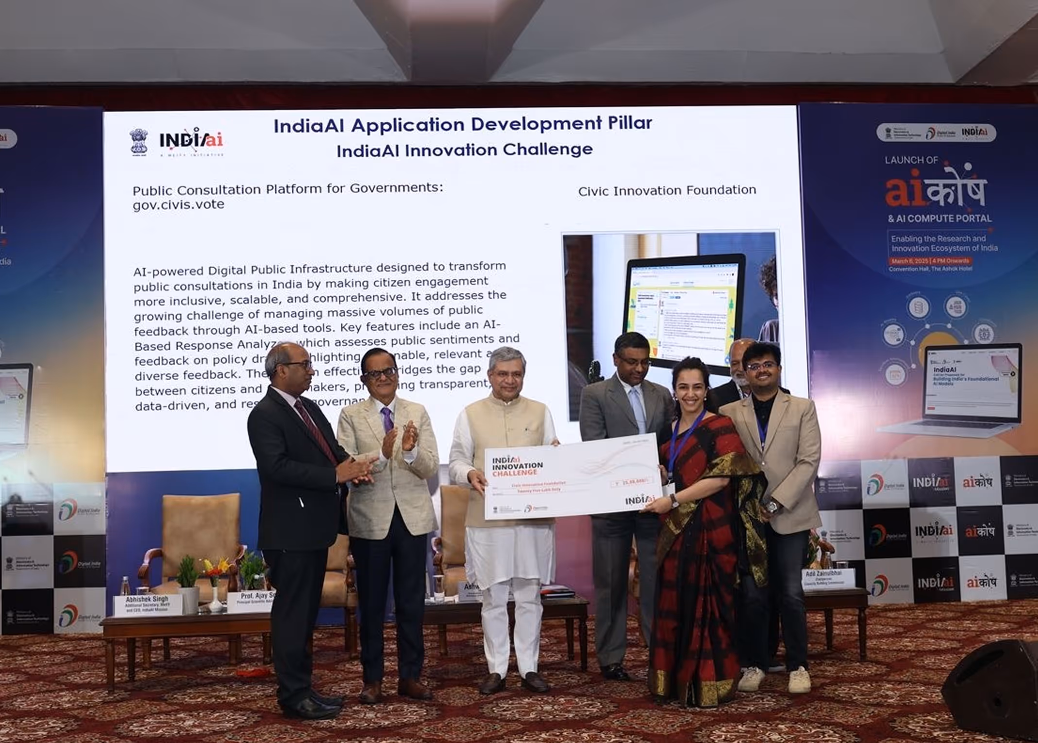 Group of six people standing on stage at the IndiaAI Innovation Challenge event holding a large ceremonial check, with a presentation slide about AI-powered public consultation platform in the background.