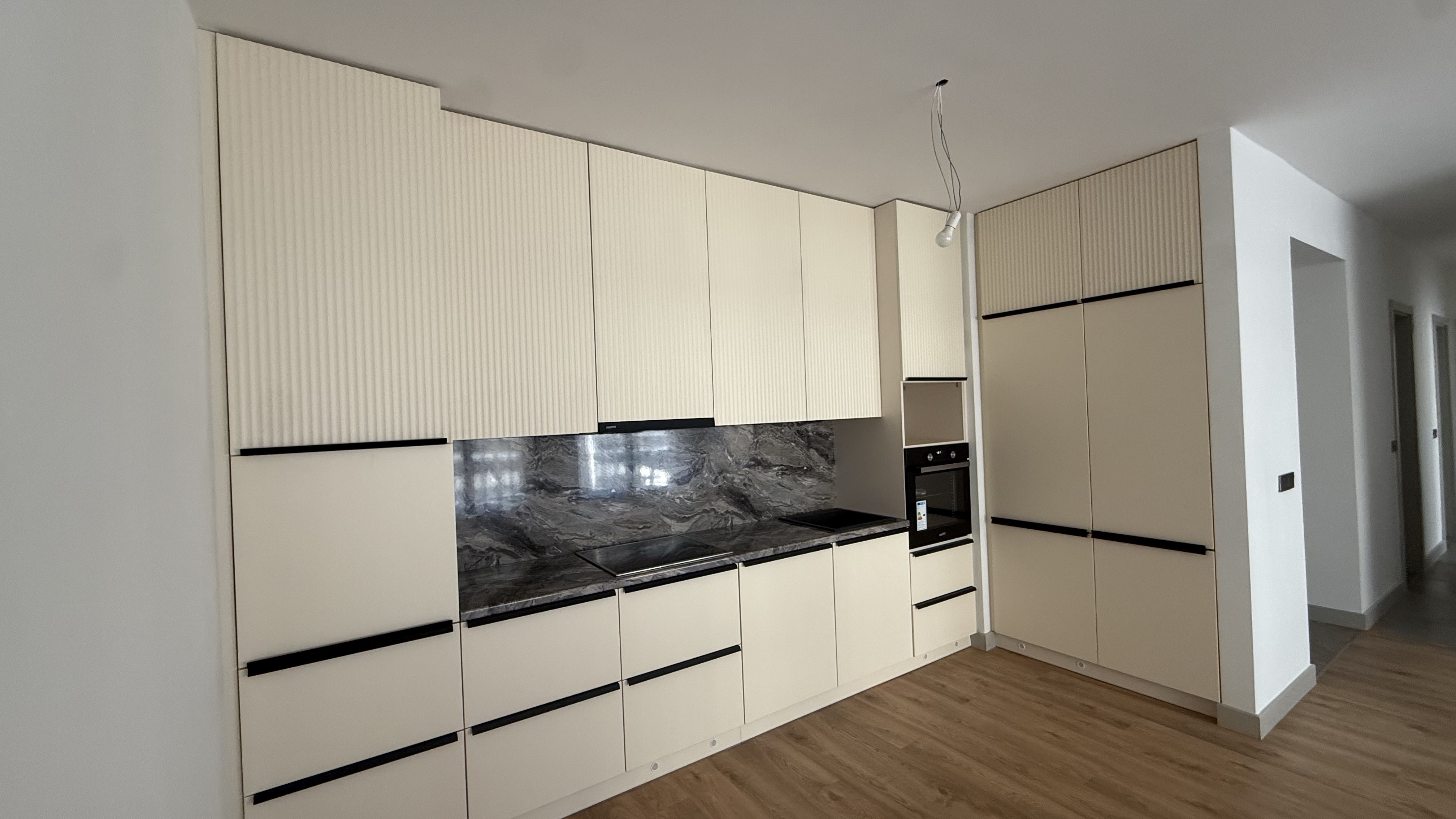 Modern kitchen with beige cabinets, marble backsplash, built-in oven, and a green sofa on wooden floor.