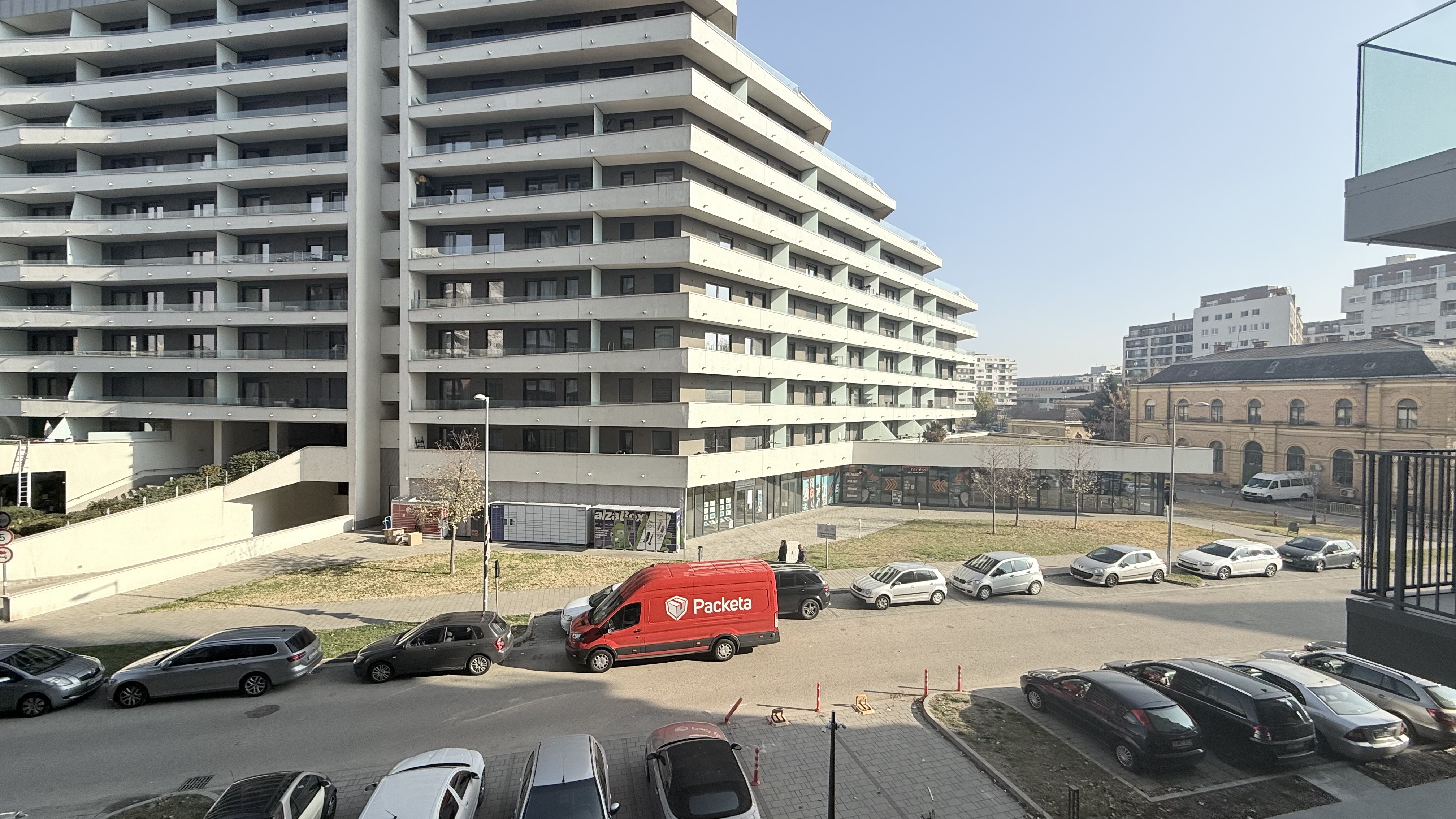 Street view of a modern multi-story apartment building with parked cars along the road, including a prominent red delivery van with 'Packeta' branding.