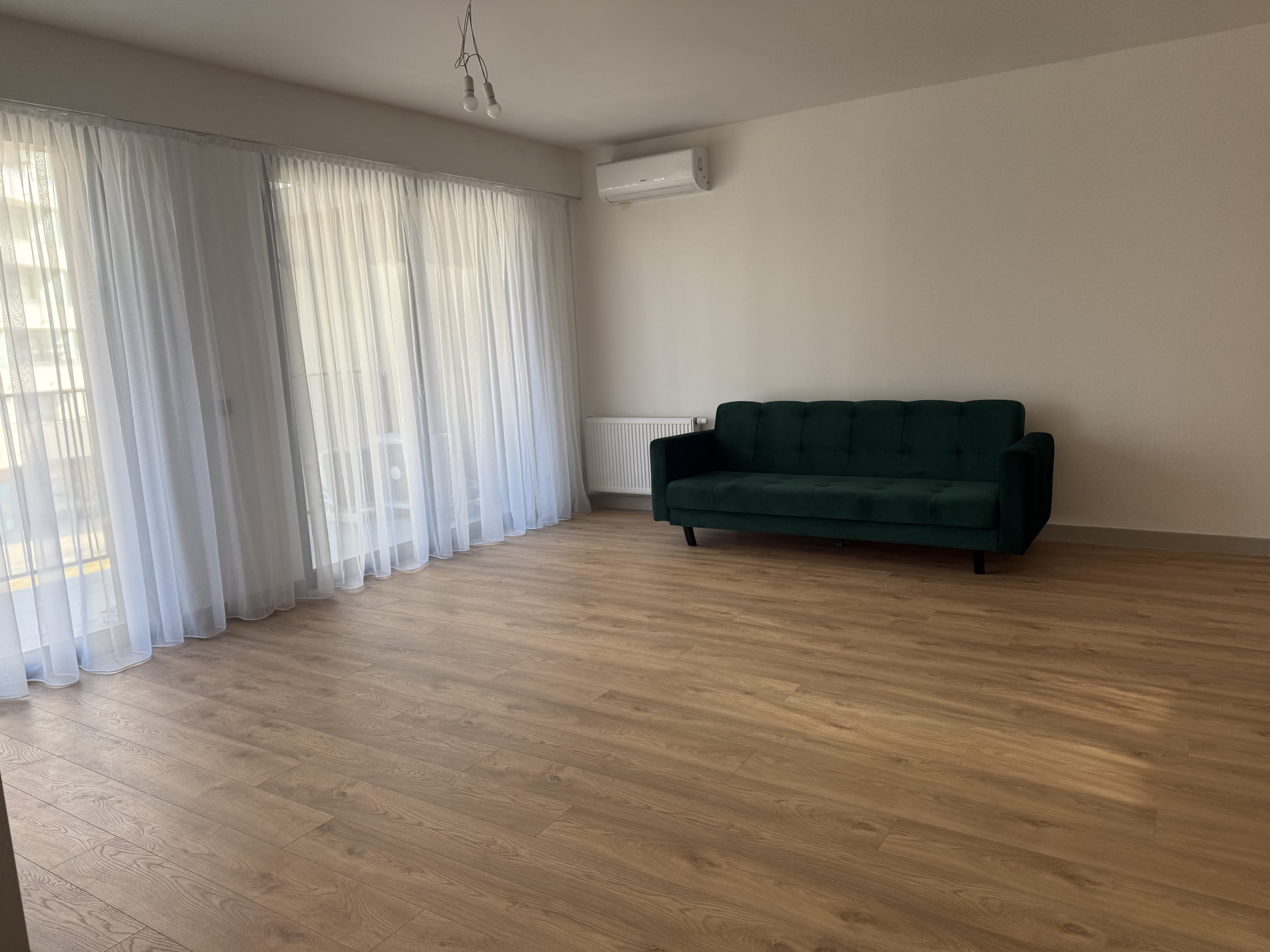 Minimalist room with light wood flooring, sheer white curtains over large windows, and a dark green sofa against a white wall.
