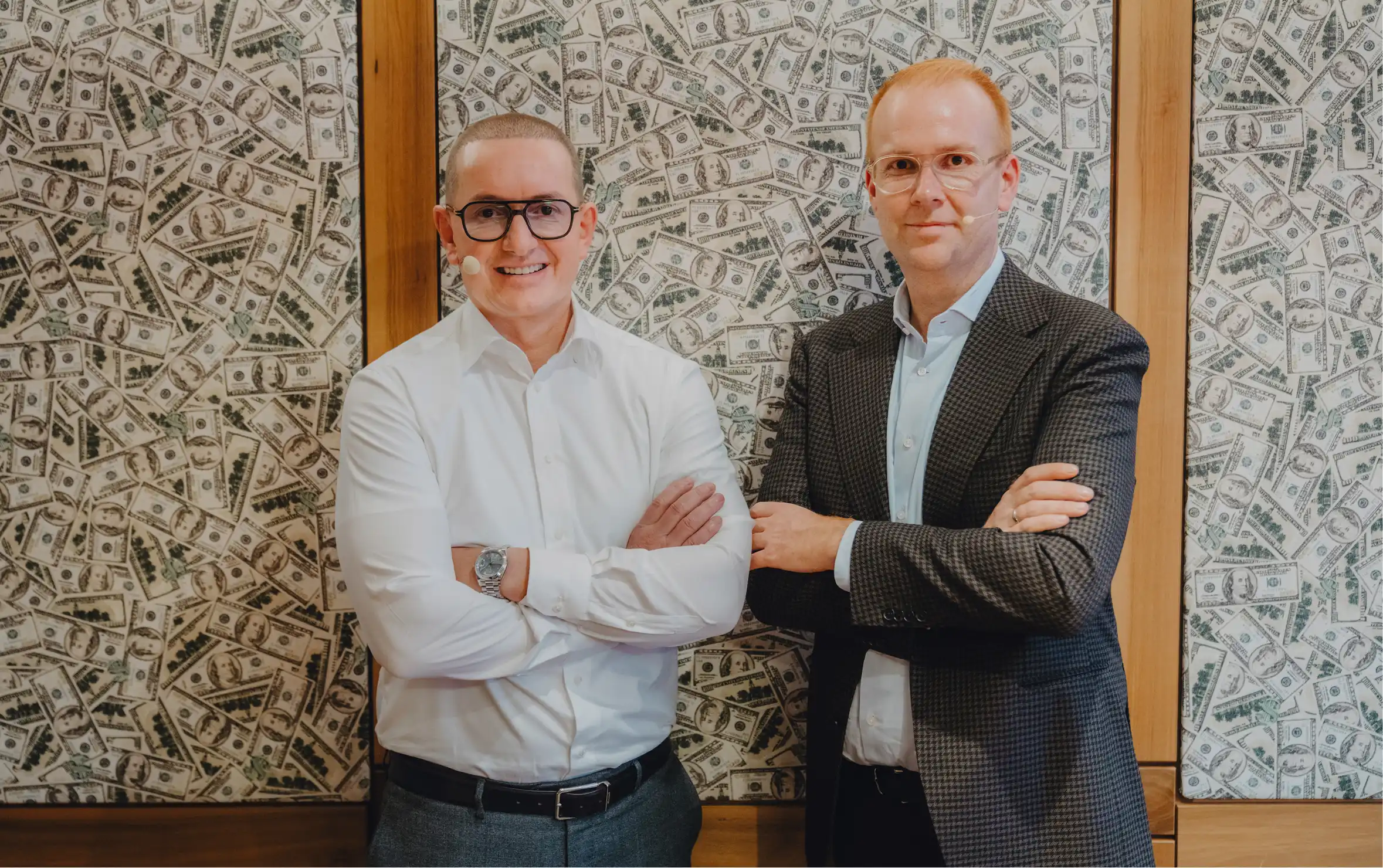 Zwei Männer mit Brille und Mikrofon stehen vor einer Wand mit Dollar-Scheinen, die Arme verschränkt.