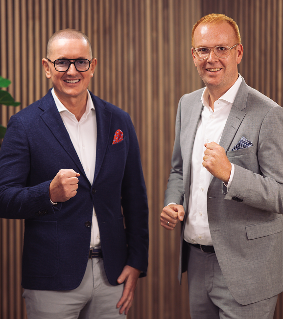 Zwei lächelnde Männer in Anzügen mit Faust geballt, vor gestreiftem Holzpaneel-Hintergrund.