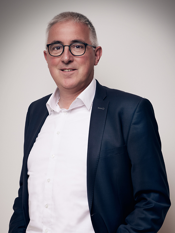 Mann mit kurzen grauen Haaren, Brille, weißem Hemd und dunkelblauem Sakko vor neutralem Hintergrund.