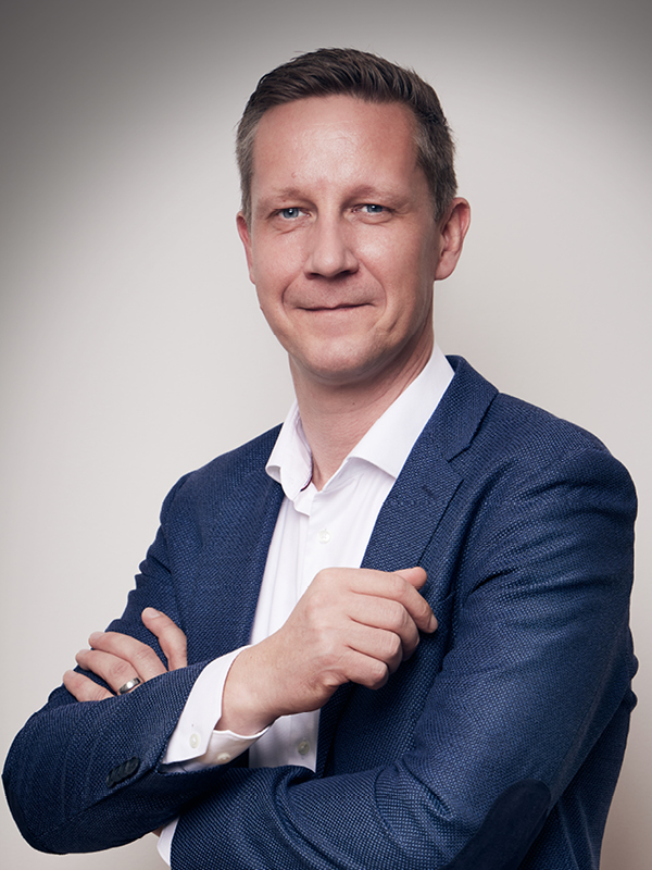 Mann mit kurzem braunem Haar, der einen blauen Blazer und ein weißes Hemd trägt, mit verschränkten Armen und einem leichten Lächeln vor hellgrauem Hintergrund.