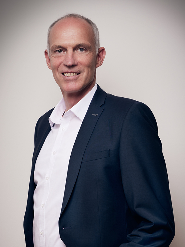 Älterer Mann mit kurzem grauen Haar, in weißem Hemd und dunkelblauem Anzug, lächelt vor hellem Hintergrund.