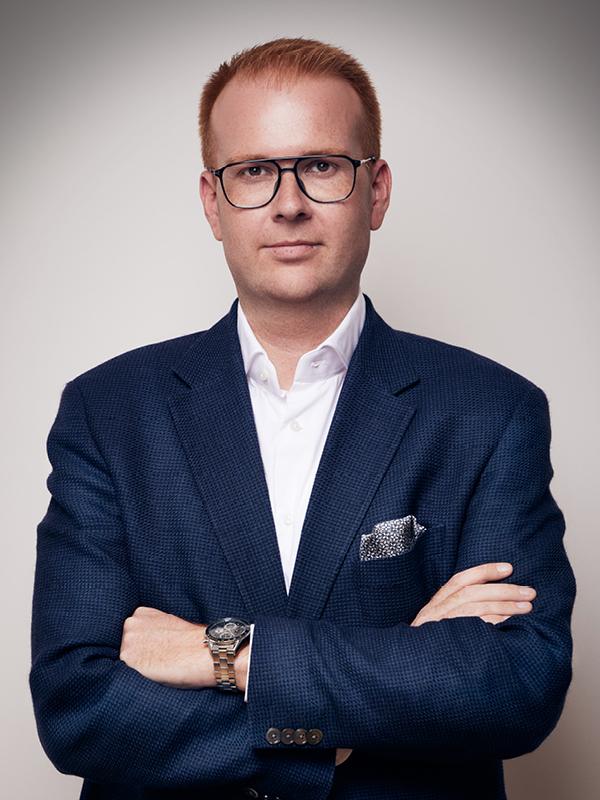 Porträt eines Mannes mit roten Haaren und Brille, der einen dunklen Anzug und ein weißes Hemd trägt, mit verschränkten Armen vor neutralem Hintergrund.