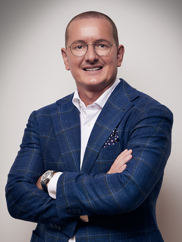 Ein Mann mit kurzen Haaren, der eine Brille trägt und in einem dunkelblauen karierten Sakko und weißem Hemd mit verschränkten Armen lächelt.