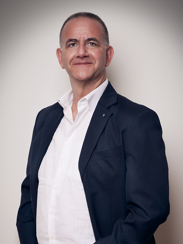 Porträt eines lächelnden Mannes mit kurzem Haar in weißem Hemd und dunklem Sakko vor hellem Hintergrund.