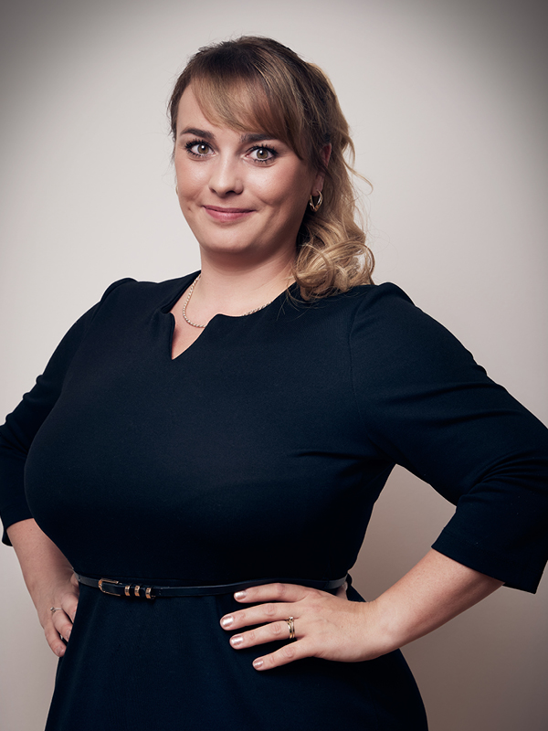 Lächelnde Frau mit blondem Haar, das zu einem lockeren Pferdeschwanz gebunden ist, trägt ein schwarzes Kleid mit Gürtel und steht vor hellem Hintergrund.