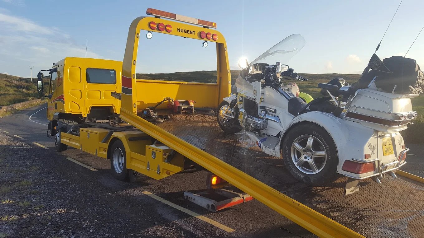 Yellow flatbed tow truck carrying a white three-wheeled motorcycle on a rural road.