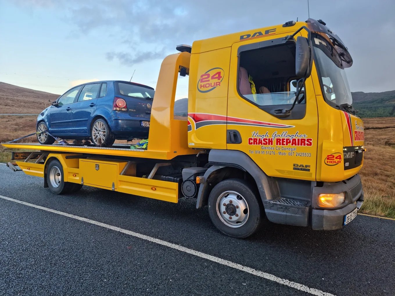 Yellow tow truck carrying a blue Volkswagen Polo on a rural road near hills.