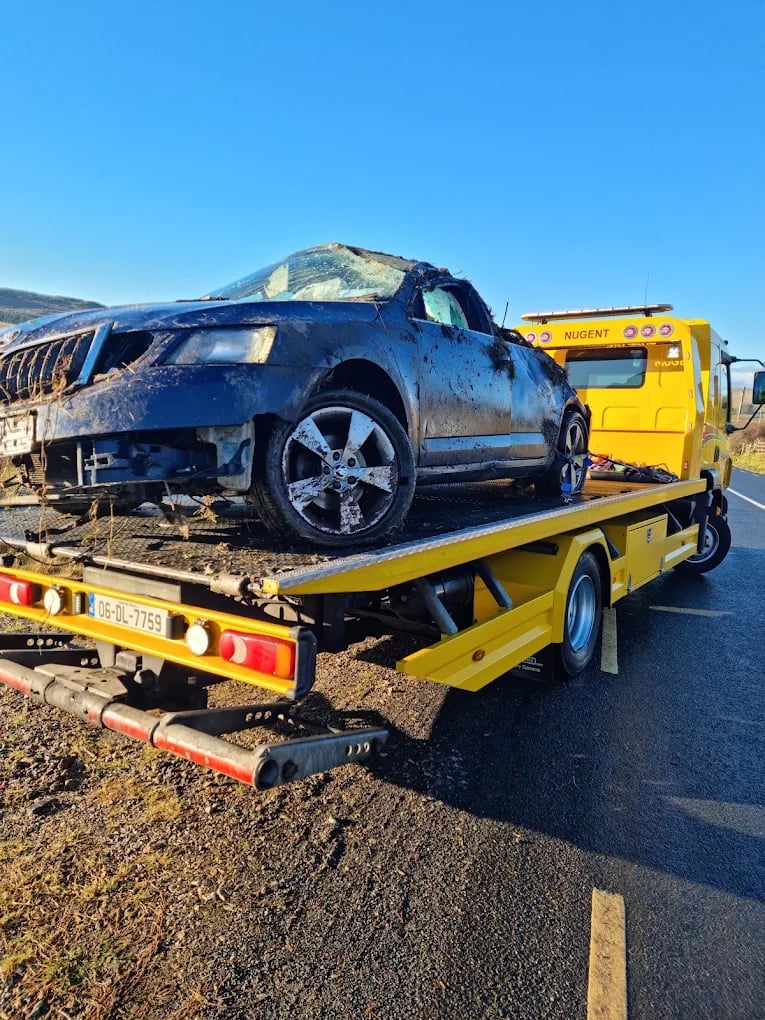 Damaged black car covered in mud on the back of a yellow tow truck on a rural road under clear blue sky.