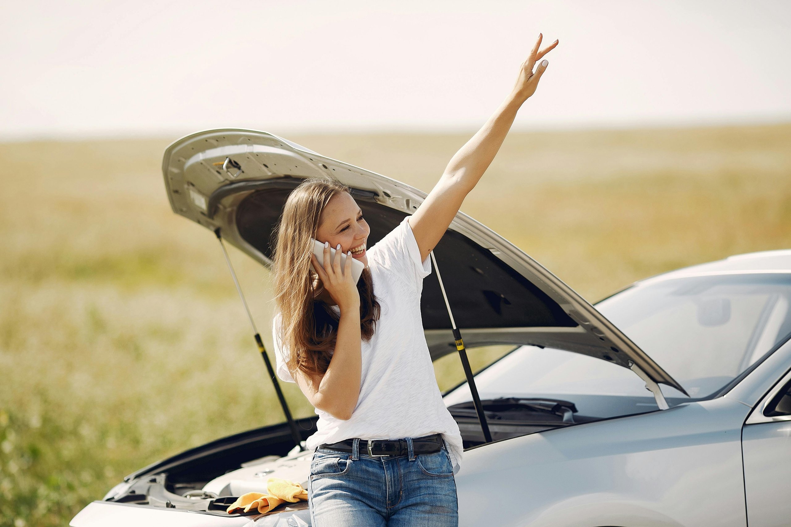 Smiling woman on phone standing beside a silver car with its hood open in a field.
