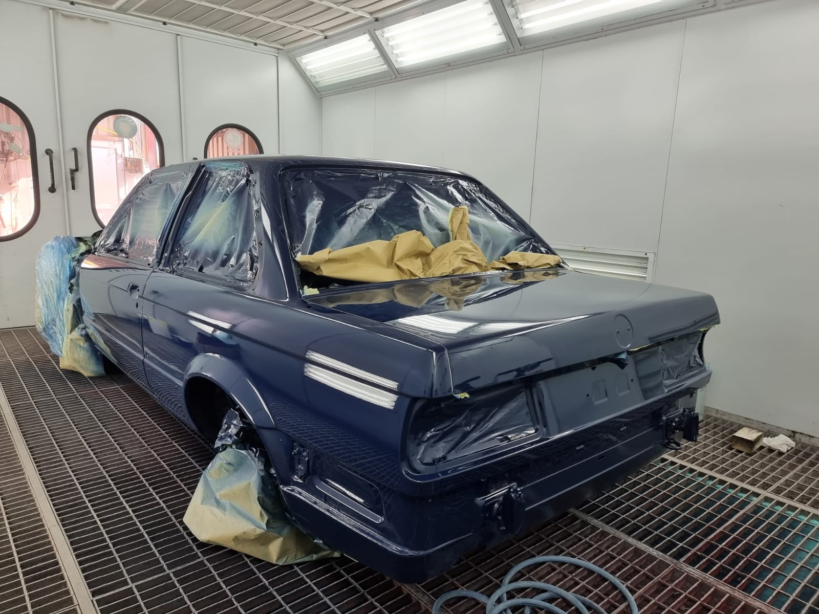 Dark blue car inside a paint booth partially masked with paper and plastic for repainting.