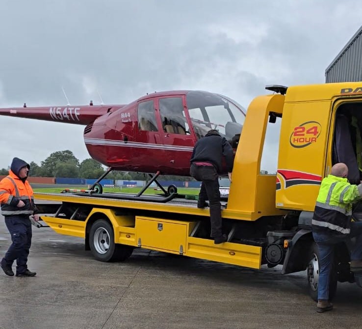 Red Robinson R44 helicopter being loaded onto a yellow 24-hour flatbed tow truck by three workers in safety jackets.