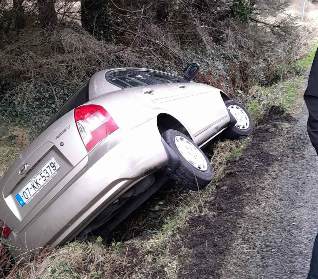 Beige Hyundai sedan stuck in a roadside ditch next to a muddy path with trees in the background.