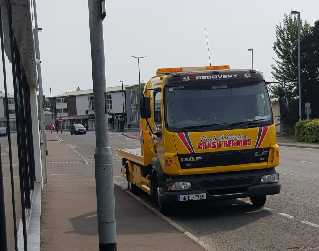 Yellow recovery truck parked on the side of a quiet urban street with buildings and a few cars in the background.