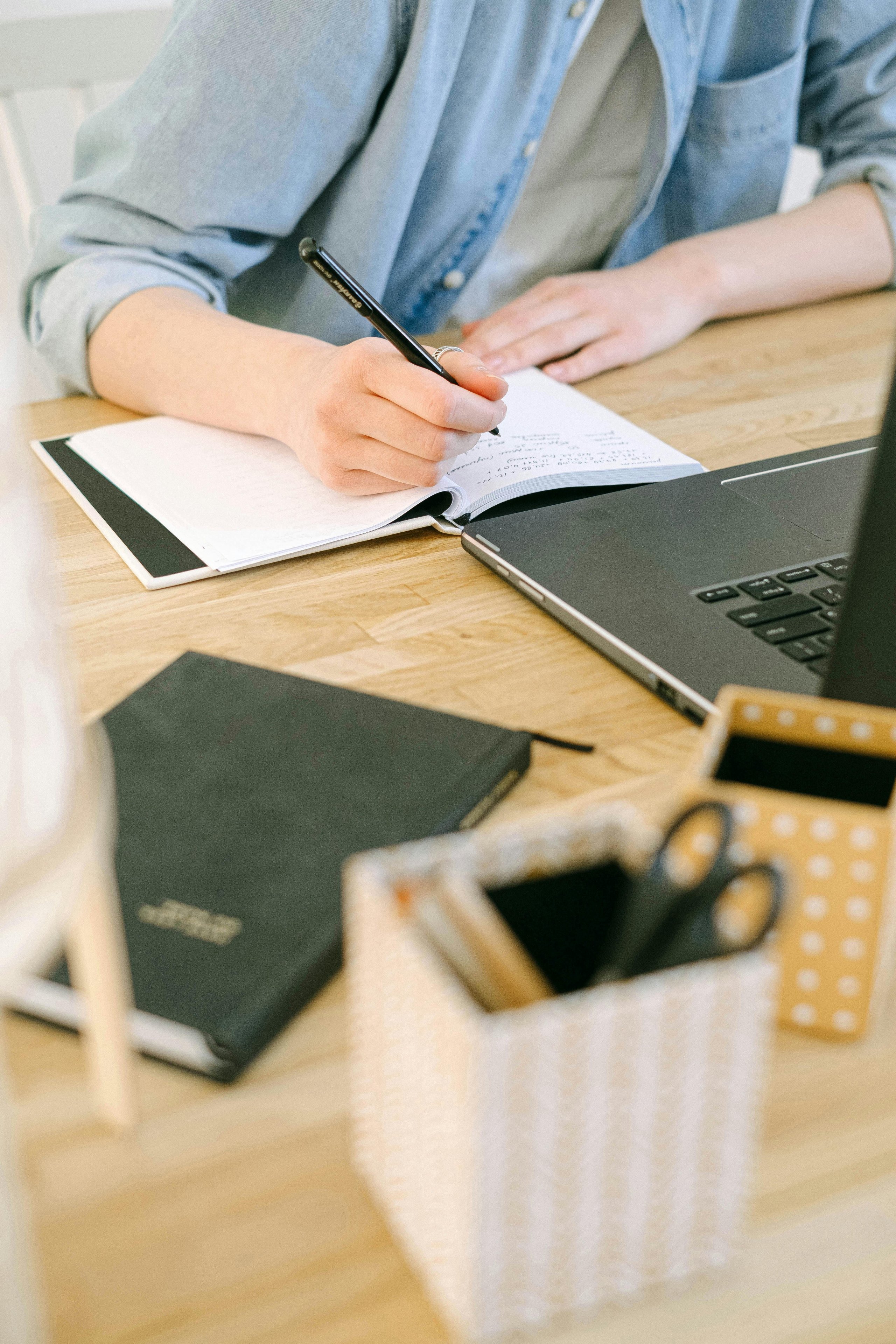 Person in a denim shirt writing notes in a notebook next to a laptop on a wooden desk with stationery items.