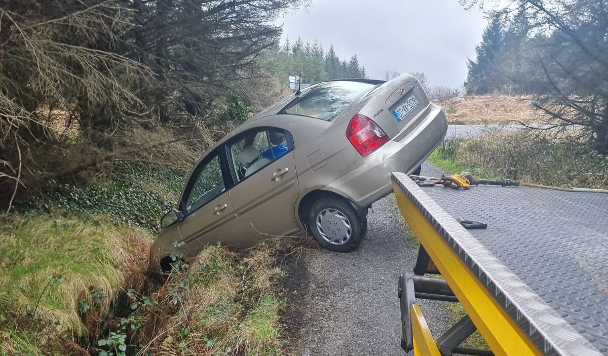 A beige sedan car partially stuck in a roadside ditch with its rear lifted onto the flatbed of a yellow tow truck.