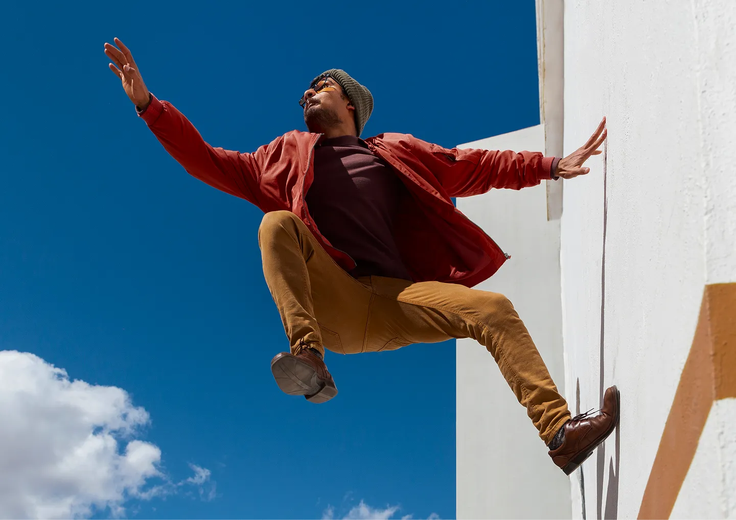 Man wearing sunglasses, a beanie, and a red jacket jumping against a white wall with a clear blue sky background.