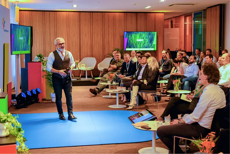 Man giving a presentation on a blue carpet in front of an audience seated with tables in a modern conference room.