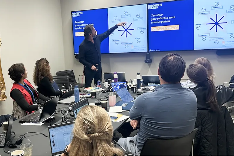 Man in a meeting room pointing at presentation slides on two large screens while colleagues watch attentively.