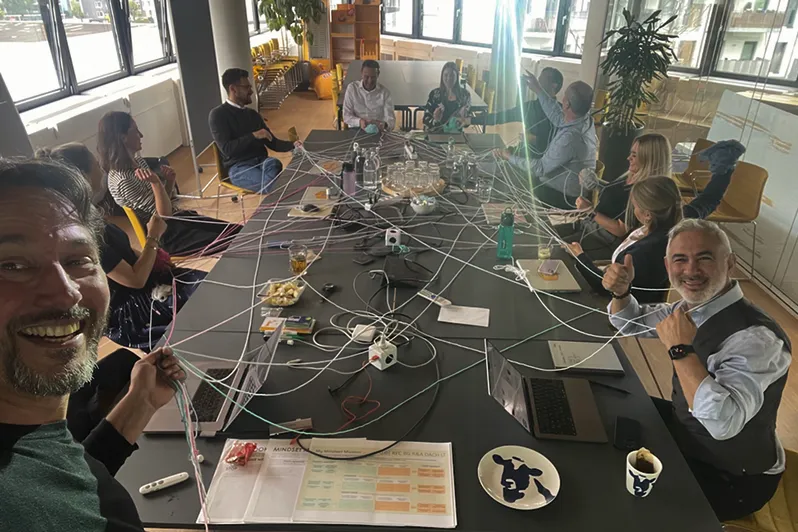 Group of people sitting around a large table in an office, holding strings connected to each other, demonstrating teamwork and collaboration.